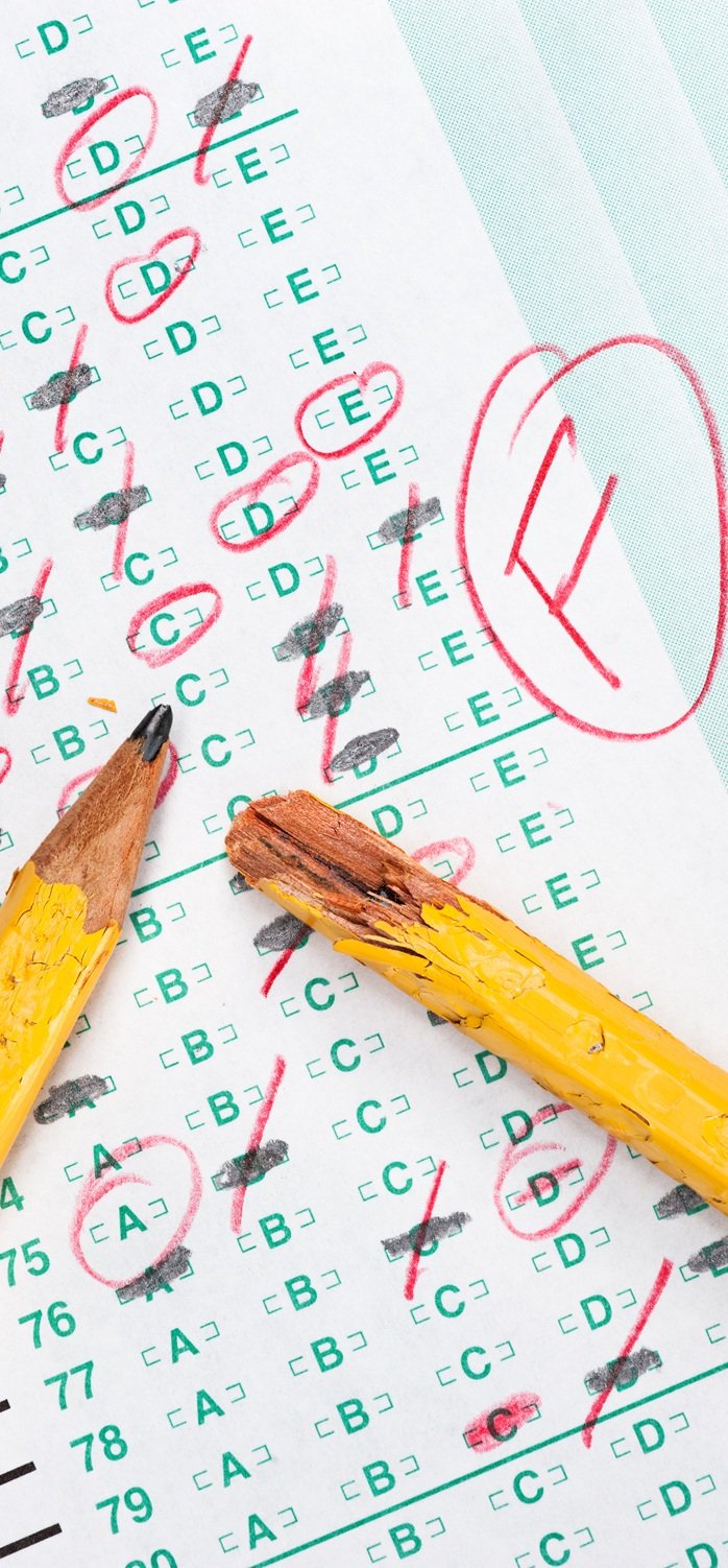 A graded test form with red scoring pencil marks indicates frustration and failure in the education system.